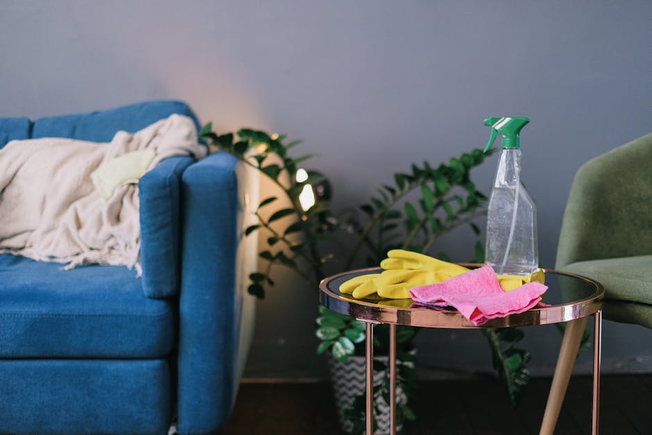 An arrangement of cleaning tools and supplies on a white surface, including a pair of pink rubber gloves positioned at the top left, a pink cleaning cloth at the top right, a green and blue spray bott
